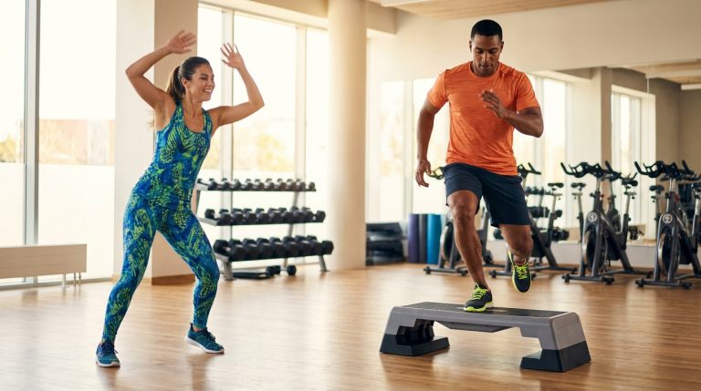 Un homme et une femme pratiquent le step et des mouvements de danse dans une salle de sport lumineuse.